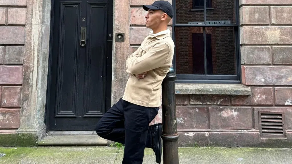 man leaning on bollard wearing black cap, beige jumper and black trousers filming Instagram content