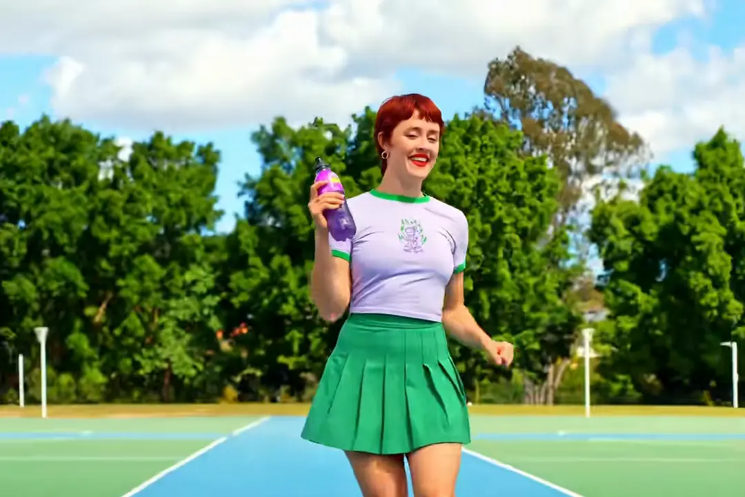 Creator @peachyfeverr in a green tennis skirt on a blue hard court holding a purple sports bottle