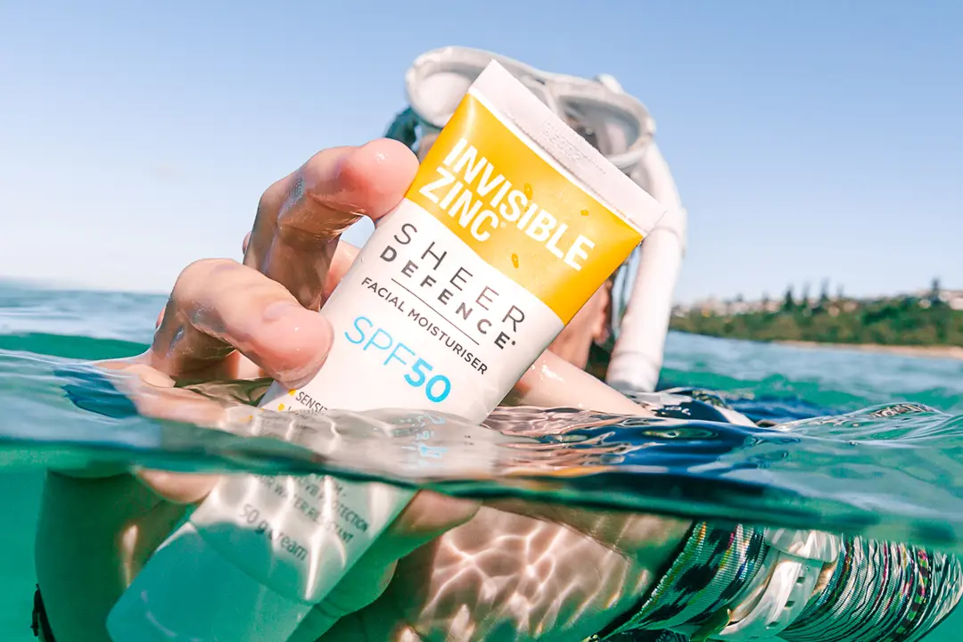 A snorkeller holding up Invisible Zinc Sheer Defence SPF50 out of turquoise water off an Australian headland, photographed by @hannahprewittphotography