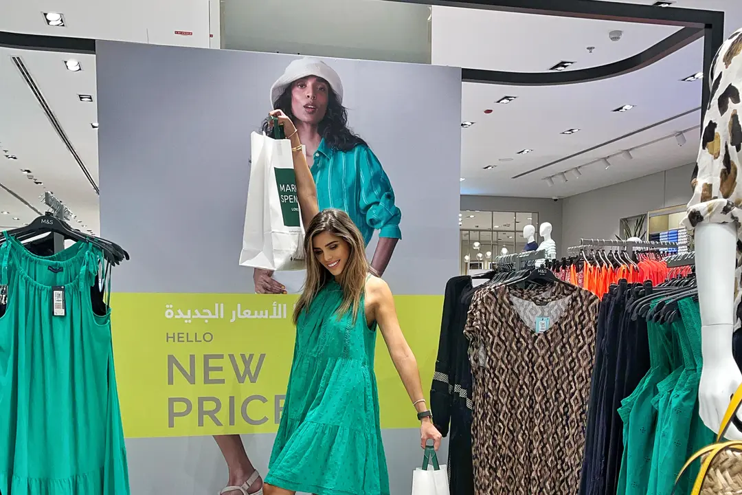Creator @danahalabii in a green dress with M&S shopping bags in front of a new-prices display in an M&S store