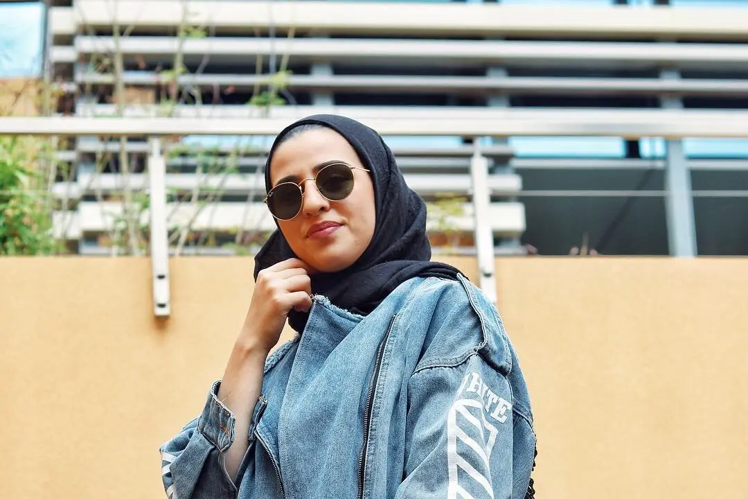 Creator @mawadda_tak in a black headscarf, round sunglasses and an oversized denim jacket, holding a red handbag, against an ochre wall
