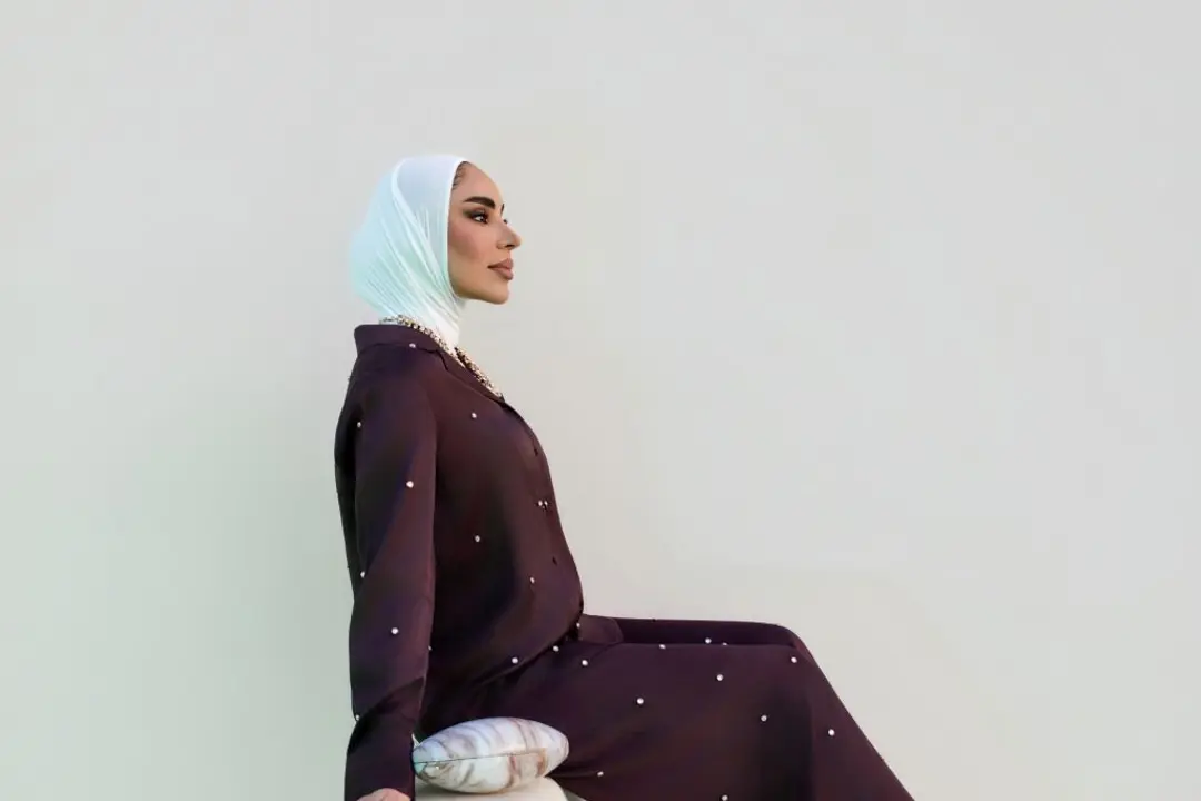 Creator @justfatema in a white headscarf and a pearl-studded plum shirt and trousers, seated in profile against a pale wall