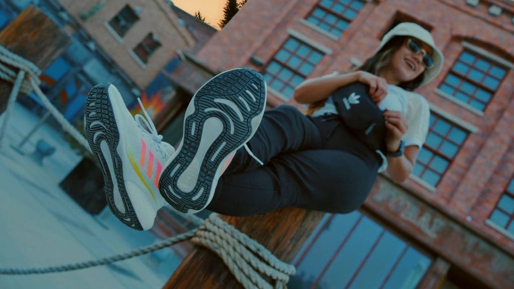 woman sitting on post wearing adidas trainers, black trousers, white top and bucket hat