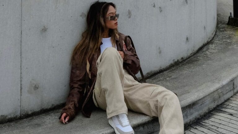 woman sitting on curb wearing brown leather jacket cream trousers and white trainers